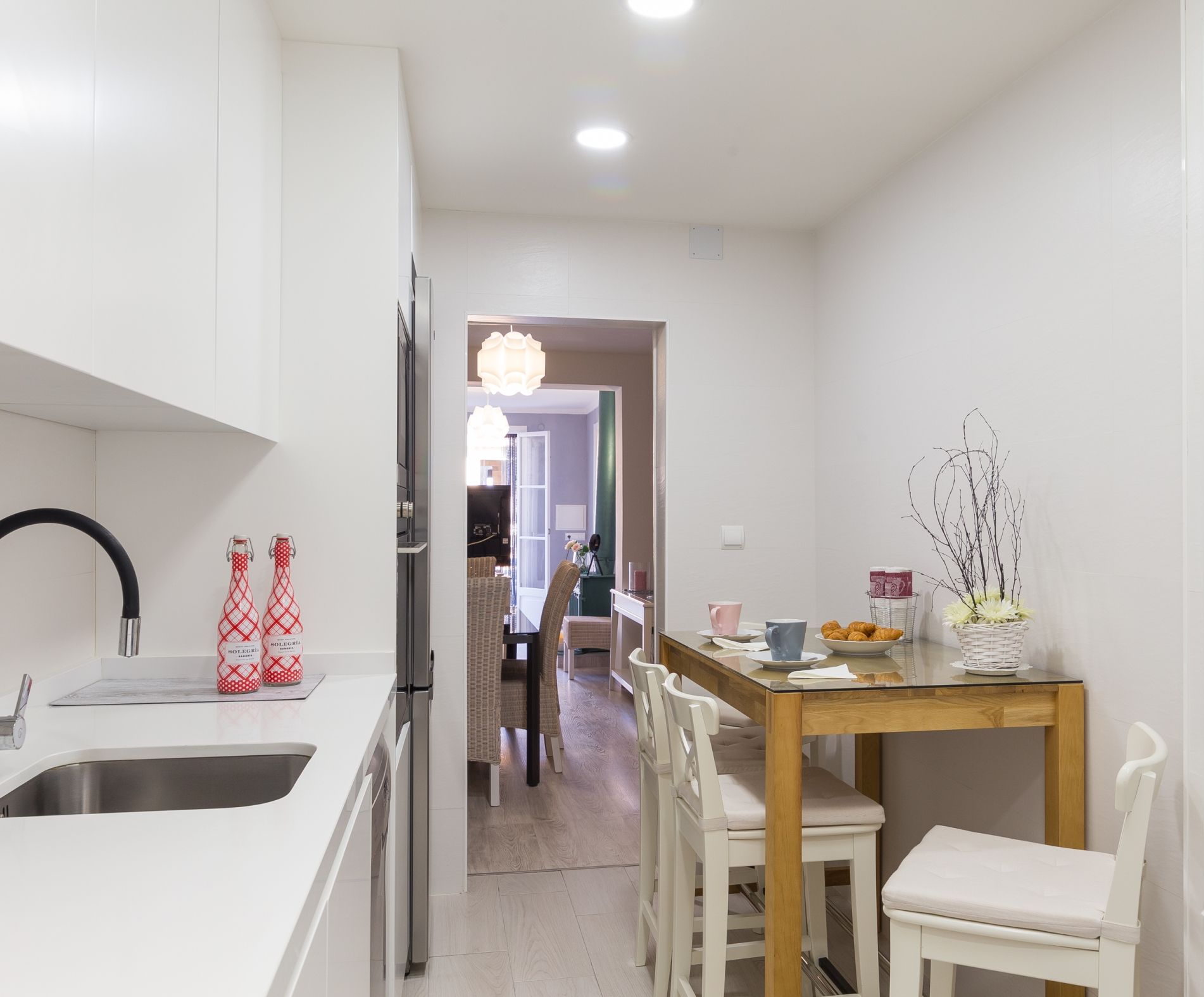 Una cocina moderna con armarios blancos, un lavamanos de acero inoxidable y un comedor con mesa de madera y sillas blancas.