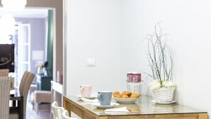 A dining area with a glass-top table, white chairs, a plate of croissants, a basket of flowers, and a cup.