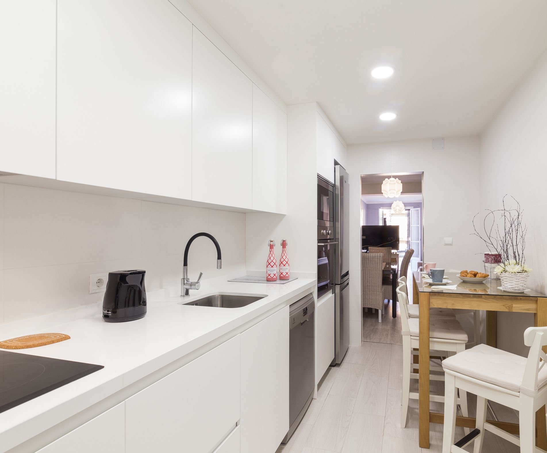Een moderne keuken met witte kastjes, een zwarte kraan en een koffiezetapparaat. Er is een zithoek met een tafel en stoelen, en een televisie op de achtergrond.