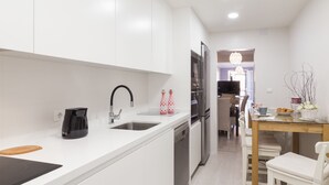 A modern kitchen with white cabinets, a black faucet, and a coffee maker. There is a dining area with a table and chairs, and a TV in the background.