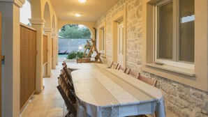 A long dining table with chairs, set in a covered outdoor area with stone walls and a window.
