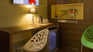 Ein Hotelzimmer mit an der Wand befestigtem Fernseher, einem Schreibtisch mit Lampe, einem Sessel, einem kleinen Kühlschrank und einem Regal mit Dekoration.
