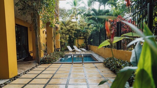 A tiled pool area with two lounge chairs, surrounded by lush greenery and a yellow building. - Casa Tres Pochotes - Beach Front Paradise (CR)