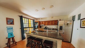 A kitchen with wooden cabinets, a stainless steel refrigerator, a water dispenser, and a bar stool area. - Casa Tres Pochotes - Beach Front Paradise (CR)