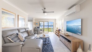 A modern living room with a sofa, coffee table, and TV. There are framed pictures on the wall and a view of the sea through the glass doors. - Sandy Beach House Corlette, 1/146 Sandy Point Road - Waterfront, WIFI, Aircon (Corlette)
