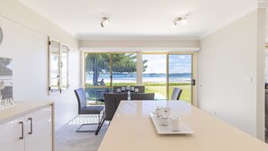 A modern dining area with a large table, chairs, and a view of the sea through large windows. - Sandy Beach House Corlette, 1/146 Sandy Point Road - Waterfront, WIFI, Aircon (Corlette)