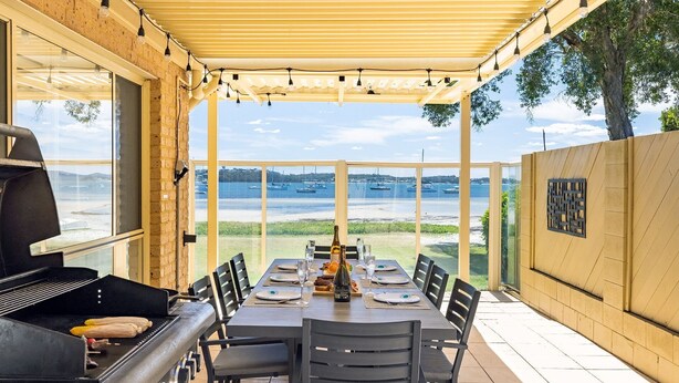 An outdoor dining area with a view of the sea and boats, a grill, and a table set for four. - Sandy Beach House Corlette, 1/146 Sandy Point Road - Waterfront, WIFI, Aircon (Corlette)