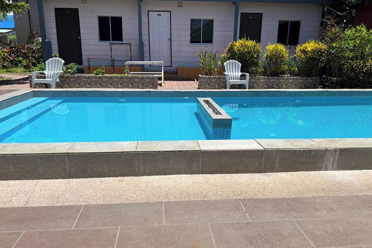 A rectangular swimming pool with a tiled deck area, two white chairs, and a small table.