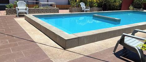 Piscina rectangular con solárium empedrado, rodeada de sillas y un banco blancos.