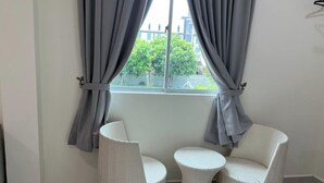 A room with two wicker chairs and a small round table, a window with grey curtains, and a view of greenery outside. - D'Lima Beach Inn (Langkawi)