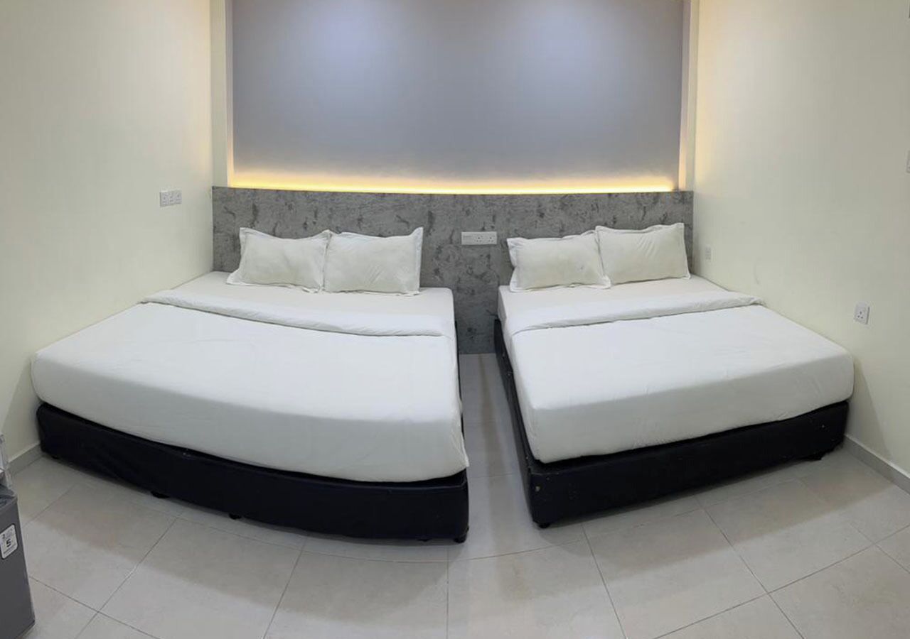 A room with two beds, each with a black frame and white bedding. The room has a modern design with a grey accent wall and a recessed lighting feature above the beds.