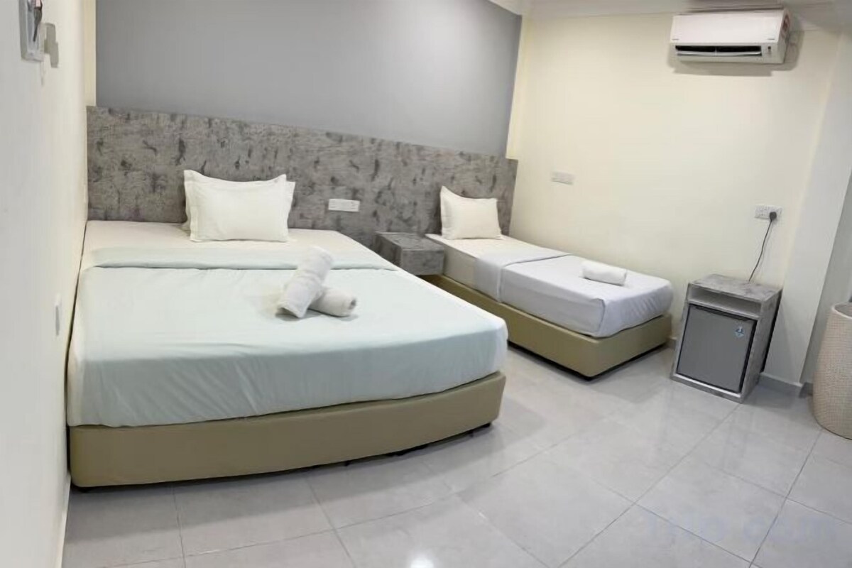 Habitación de hotel con dos camas, cabecera gris, mesita de noche pequeña y aire acondicionado instalado en la pared.