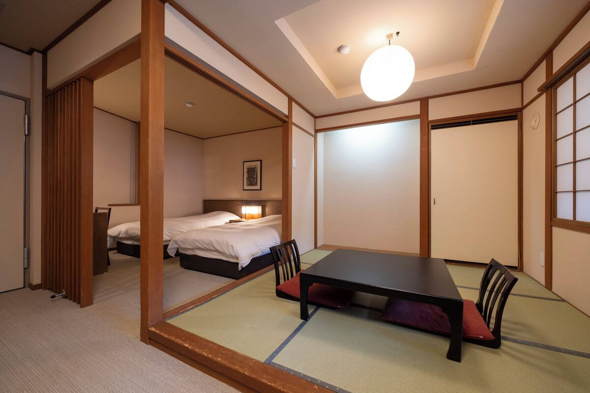 A traditional Japanese room with tatami flooring, a low table with cushions, a bed with white bedding, and sliding doors.