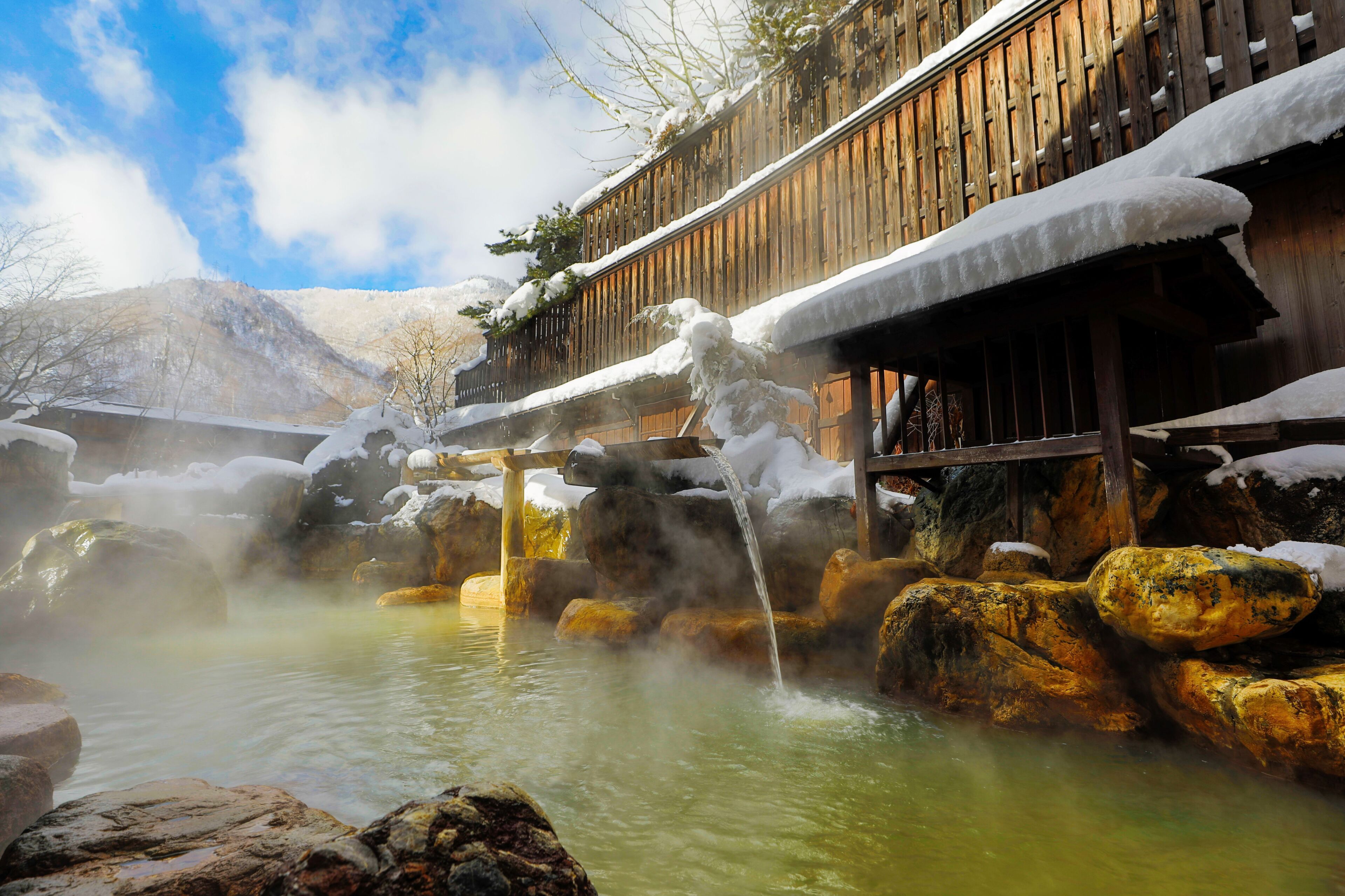 A traditional wooden building with a wooden deck, surrounded by snow-covered rocks and a hot spring area.