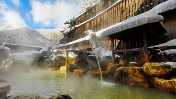 A traditional wooden building with a wooden deck, surrounded by snow-covered rocks and a hot spring area.