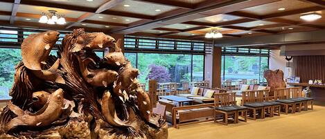 Un restaurante con una gran escultura de pescado de madera como pieza central, mesas y sillas de madera, y amplias ventanas que ofrecen vistas a la vegetación.