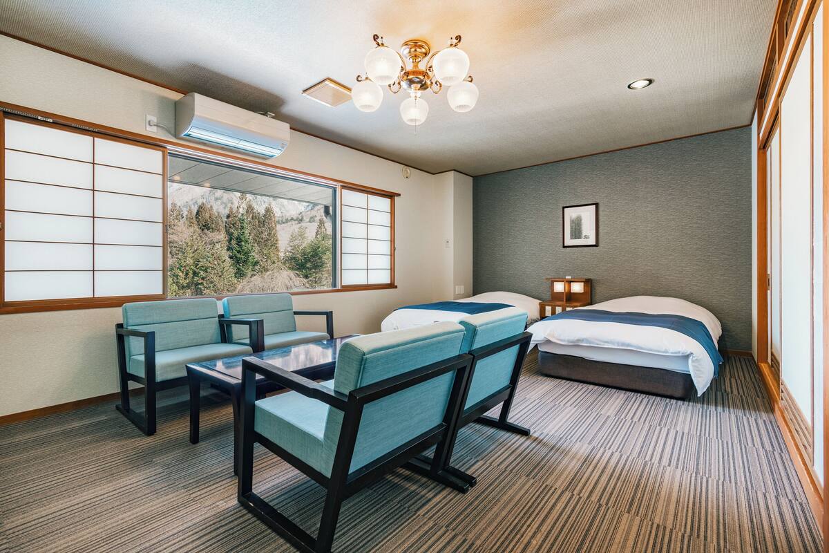 A hotel room with a large window, two beds, a table with chairs, and a ceiling light fixture.