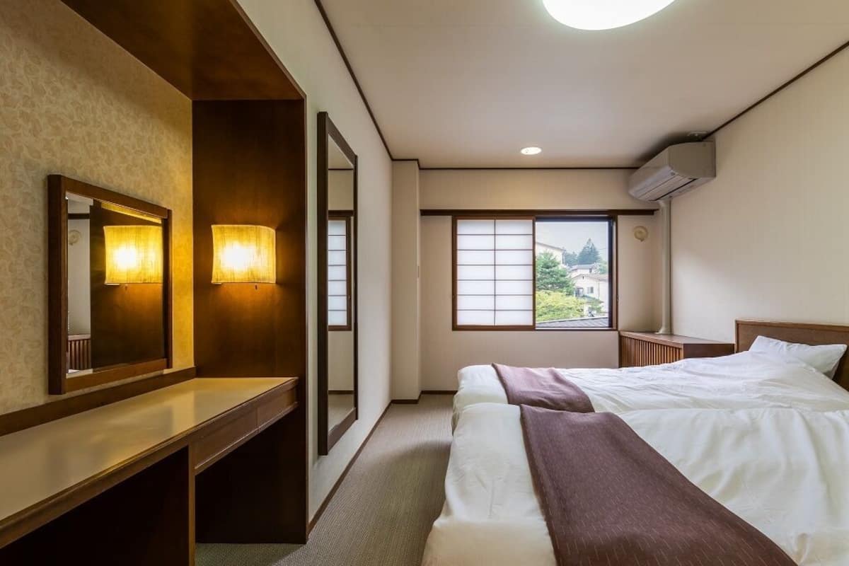 A hotel room with a bed, bedside tables, a mirror, a window with a view, and an air conditioning unit.