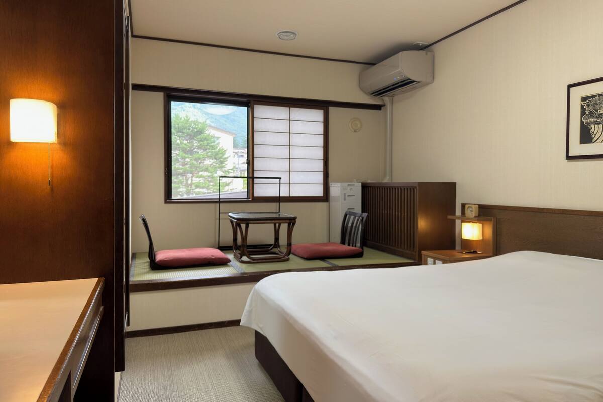 A hotel room with a bed, a desk, a chair, a window with a view, an air conditioning unit, and a wall-mounted clock.