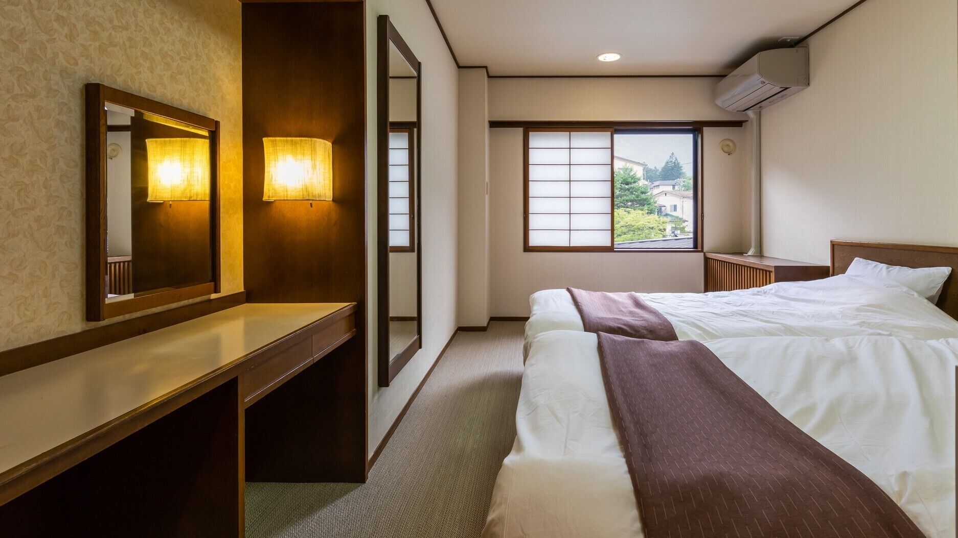 A hotel room with a bed, bedside table, mirror, wall-mounted lamp, window with view, and air conditioning unit.