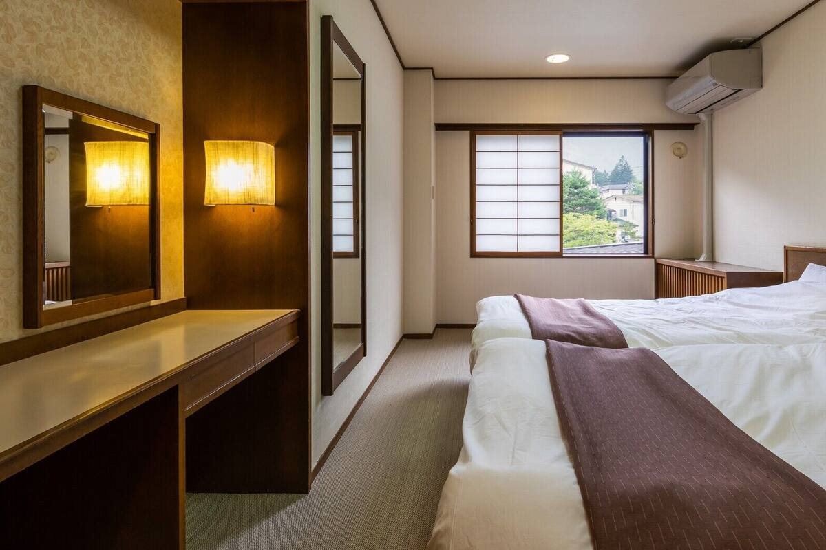 A hotel room with a bed, bedside table, mirror, wall-mounted lamp, window with view, and air conditioning unit.