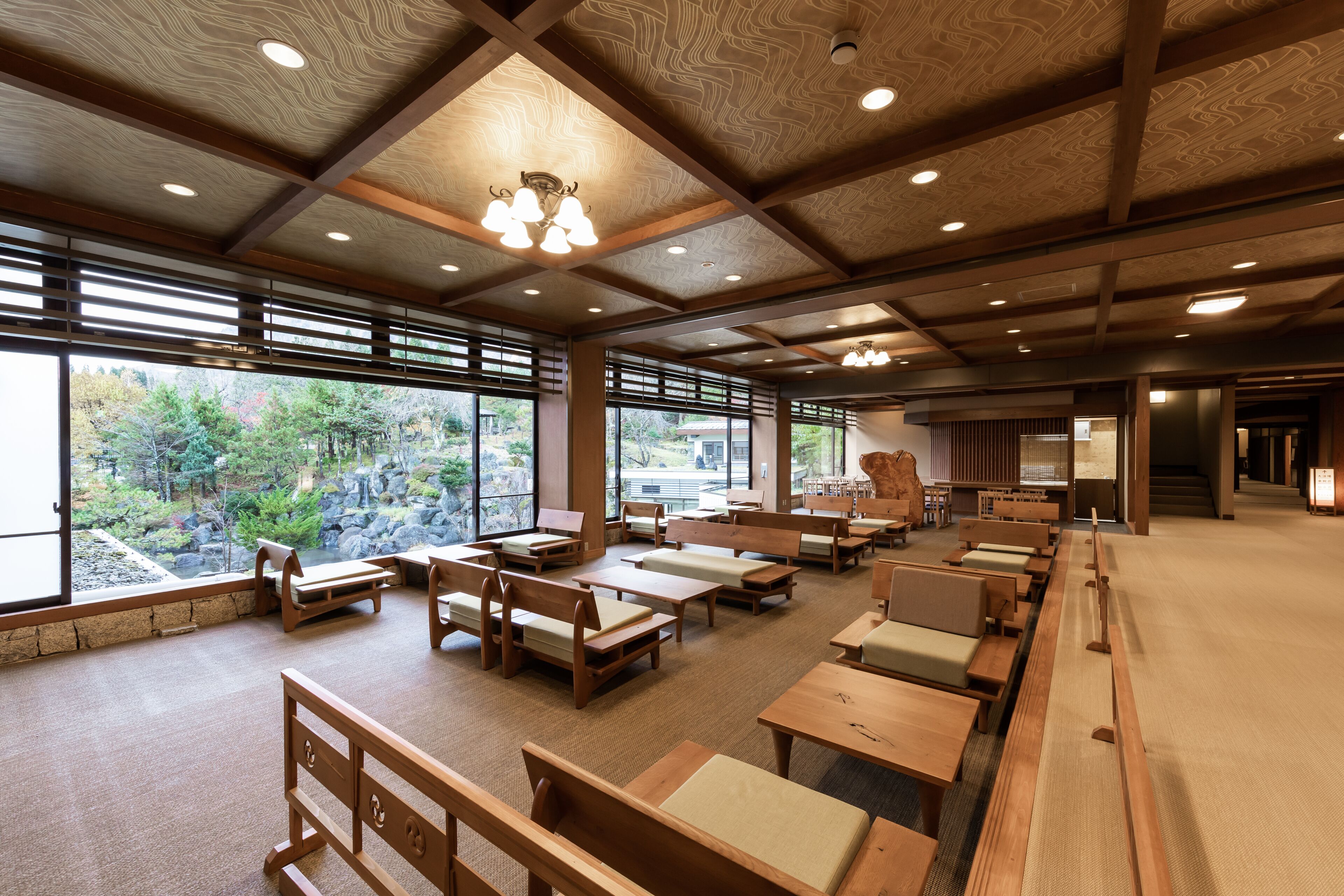 A spacious room with wooden benches and tables, large windows offering a view of a garden and trees.