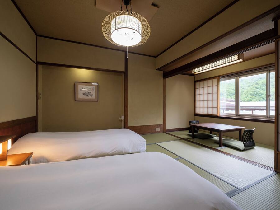 Ein traditioneller japanischer Raum mit Tatami-Boden, zwei Betten, einem niedrigen Tisch und einer Schiebetür mit Blick ins Grüne.