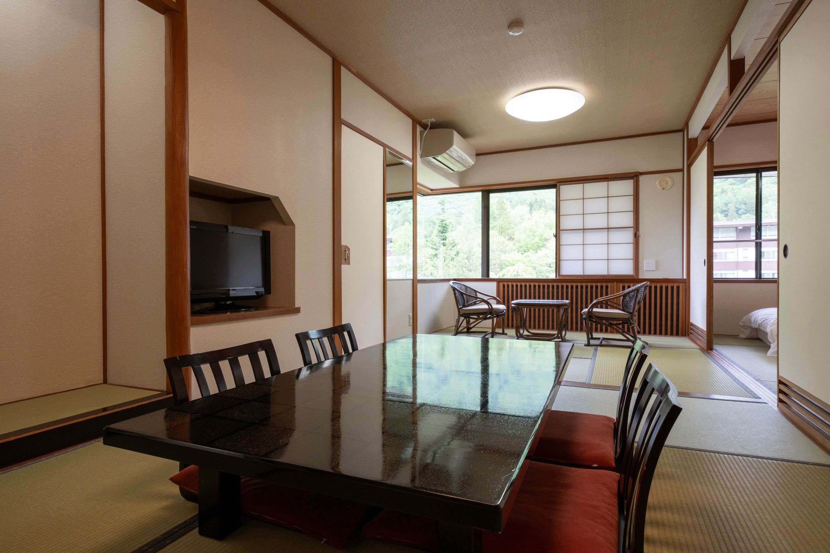 Una habitación tradicional japonesa con suelo de tatami, una mesa baja con sillas, un televisor y vistas a un bosque a través de grandes ventanales.