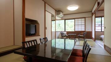 Una habitación tradicional japonesa con suelo de tatami, una mesa baja con sillas, un televisor y vistas a un bosque a través de grandes ventanales.