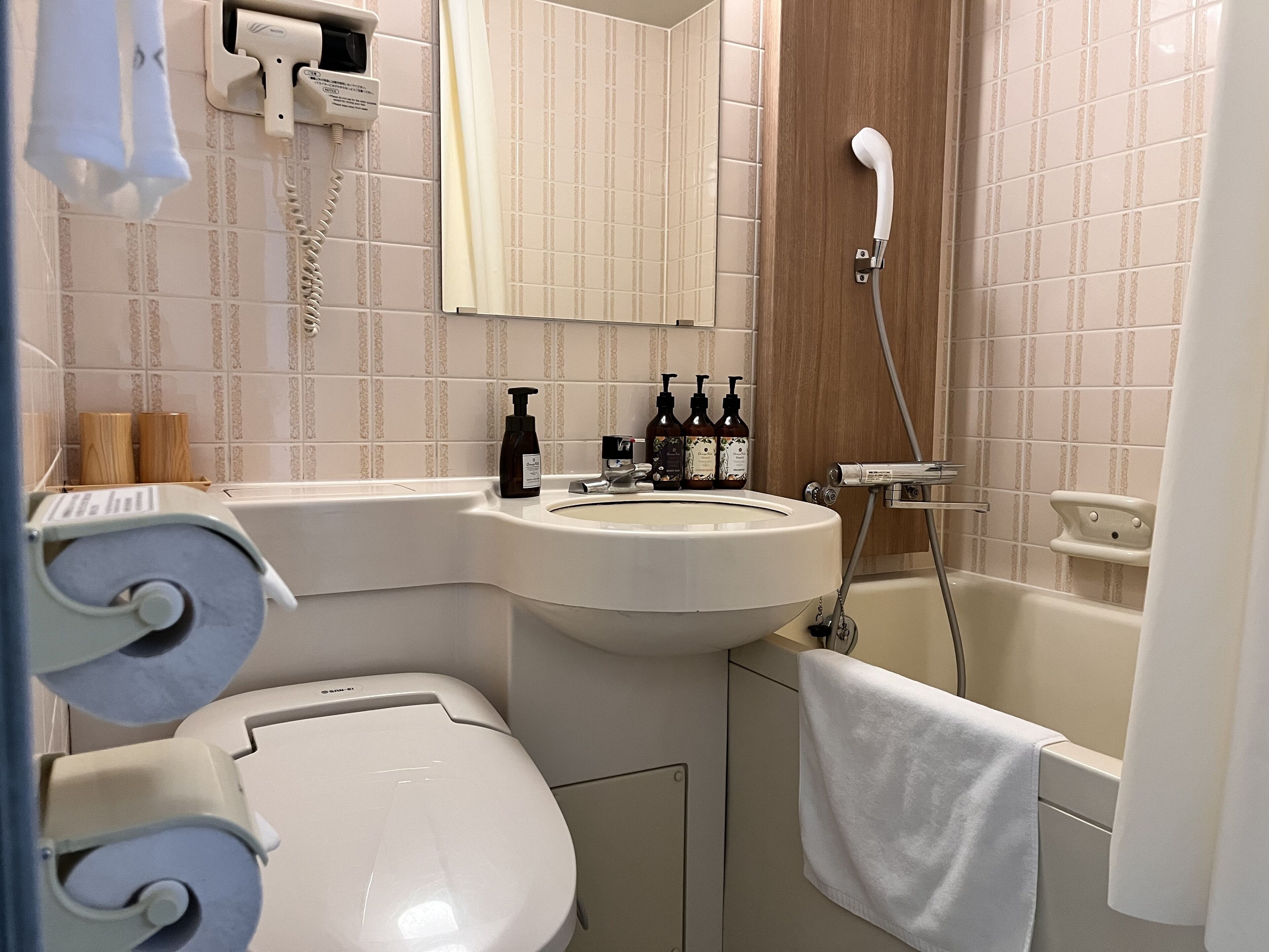 A bathroom with a toilet, sink, and shower. There are toiletries on the sink and a towel hanging on the wall.