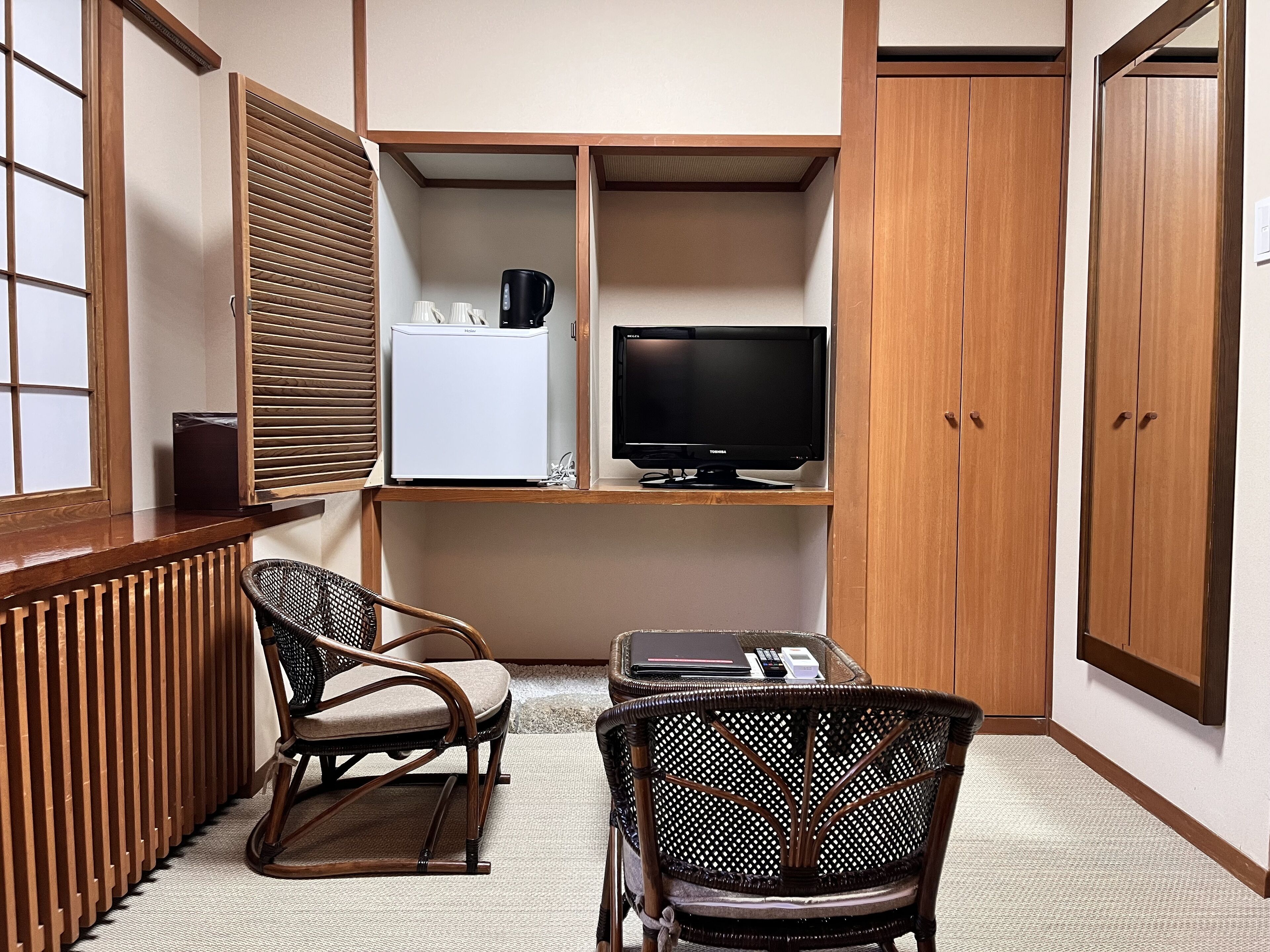 A corner room with a wooden cabinet, a television, a refrigerator, two wicker chairs, and a small table with a laptop and a phone.