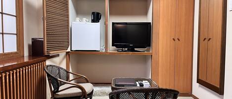 Habitación de esquina con un armario de madera, un televisor, un refrigerador, dos sillas de mimbre y una mesita con una computadora portátil y un teléfono.