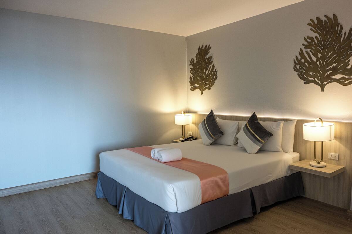 Ein ordentlich bezogenes Bett mit grau-weißer Tagesdecke, zwei Nachttischlampen und dekorativen Wandbildern.
