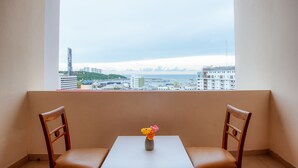 Ein Balkon mit zwei Holzstühlen, einem kleinen Tisch und einer Vase mit Blumen. Durch das Fenster ist eine Stadtansicht mit Gebäuden und einem Gewässer zu sehen.