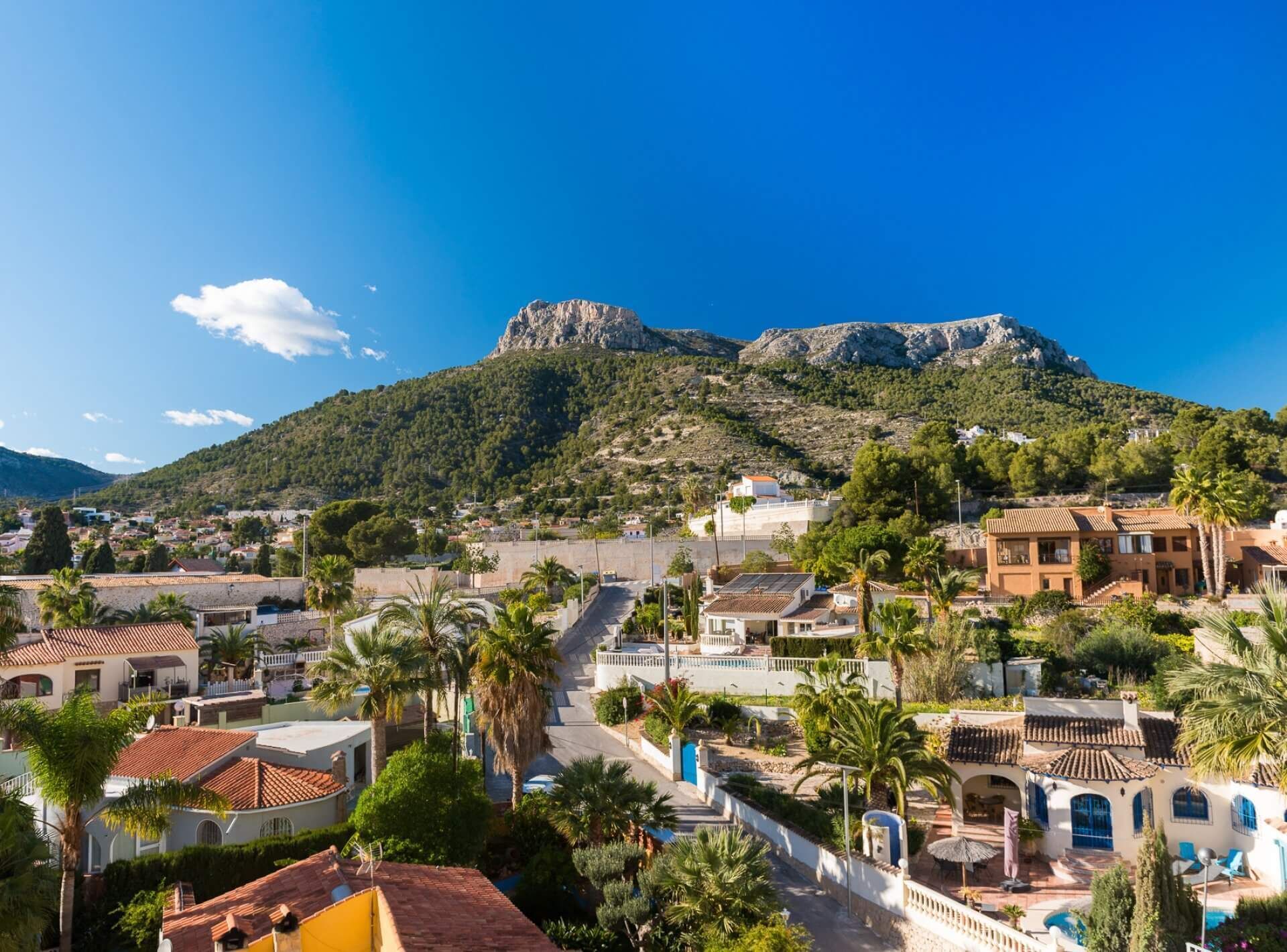 A coastal town with Mediterranean architecture, a prominent rocky hill in the background, and a clear blue sky.