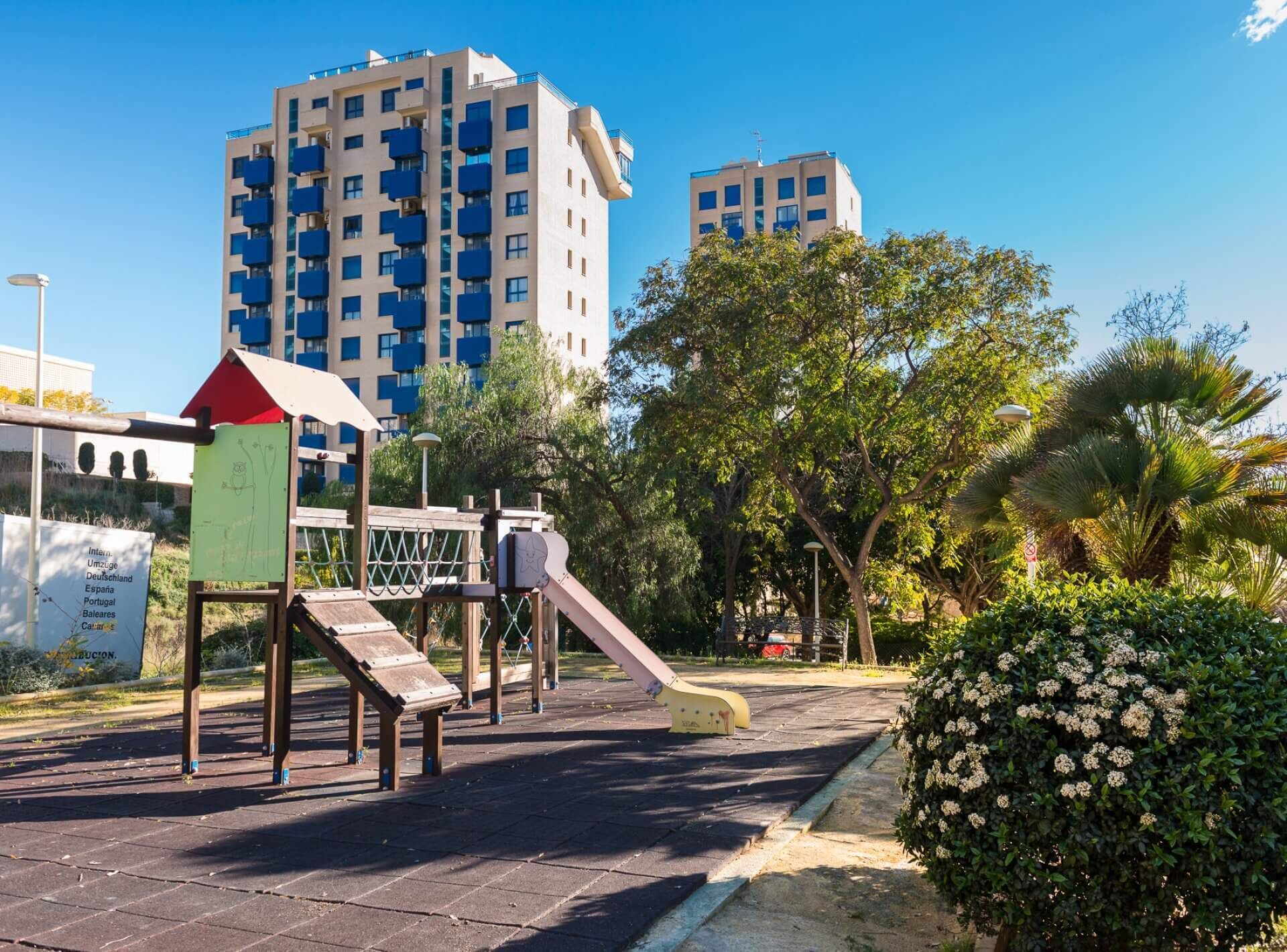 Un parc infantil amb tobogan i elements per escalar, envoltat d'arbres i un edifici al fons.