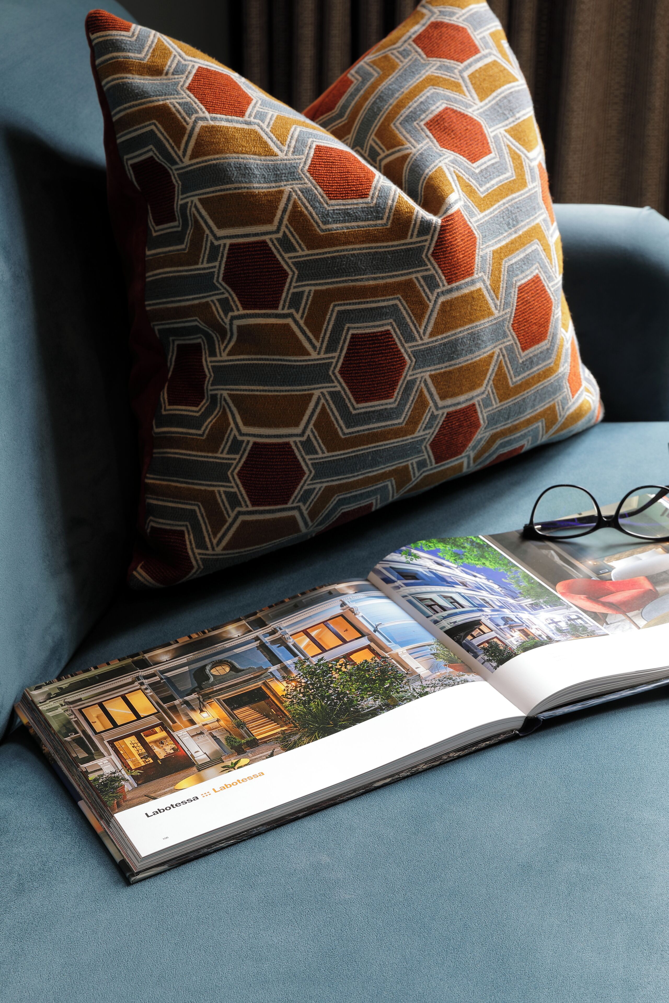 A magazine on a blue couch with a geometric patterned pillow and a pair of glasses.