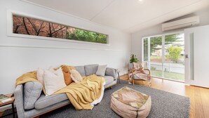 A modern living room with a grey sofa, a yellow throw blanket, and a grey rug. There is a window with a view of greenery and a brick wall. - Pet Friendly - Cutest Cottage on Fourth walk to the Beach (Rosebud)