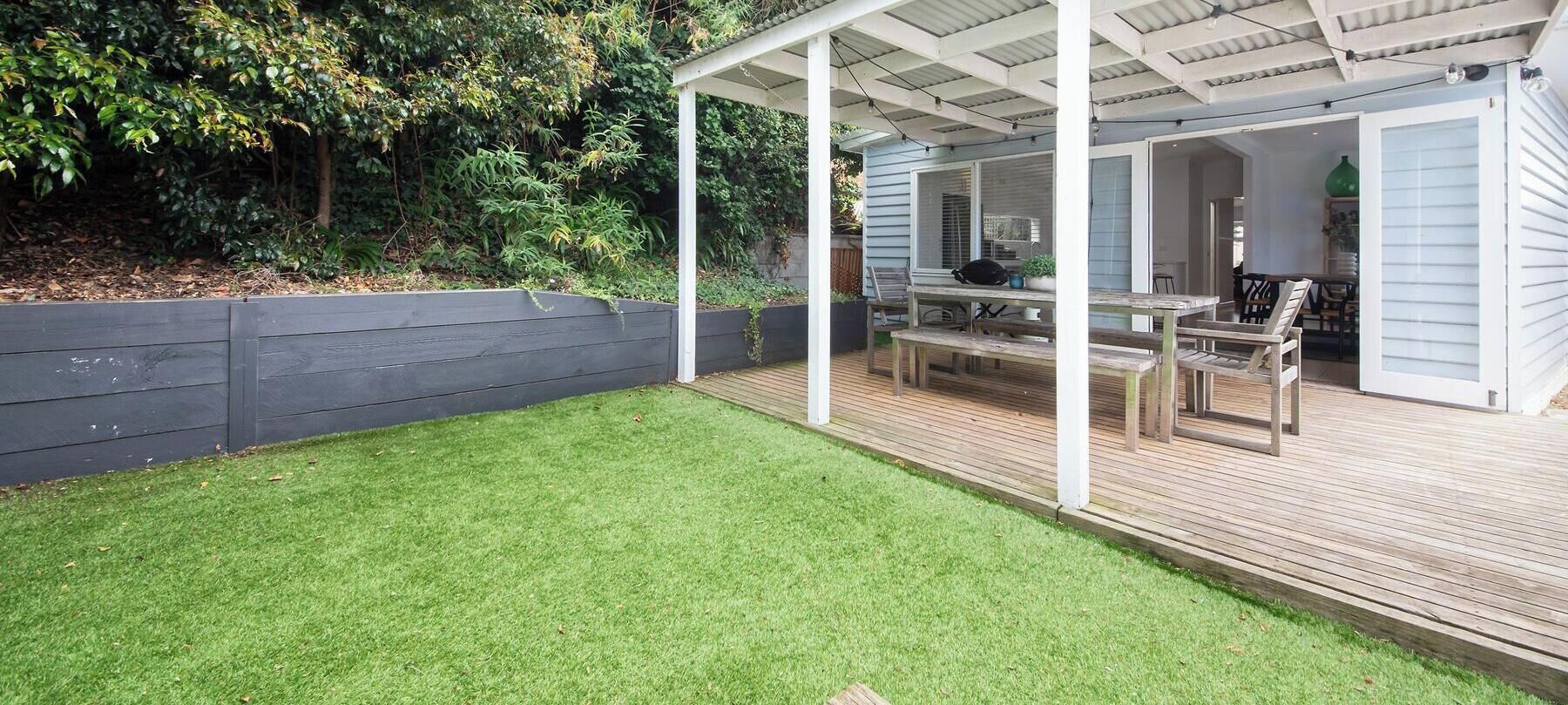 Een overdekt buitengebied met een houten terras, een tafel en stoelen, omgeven door een gazon en een houten omheining.