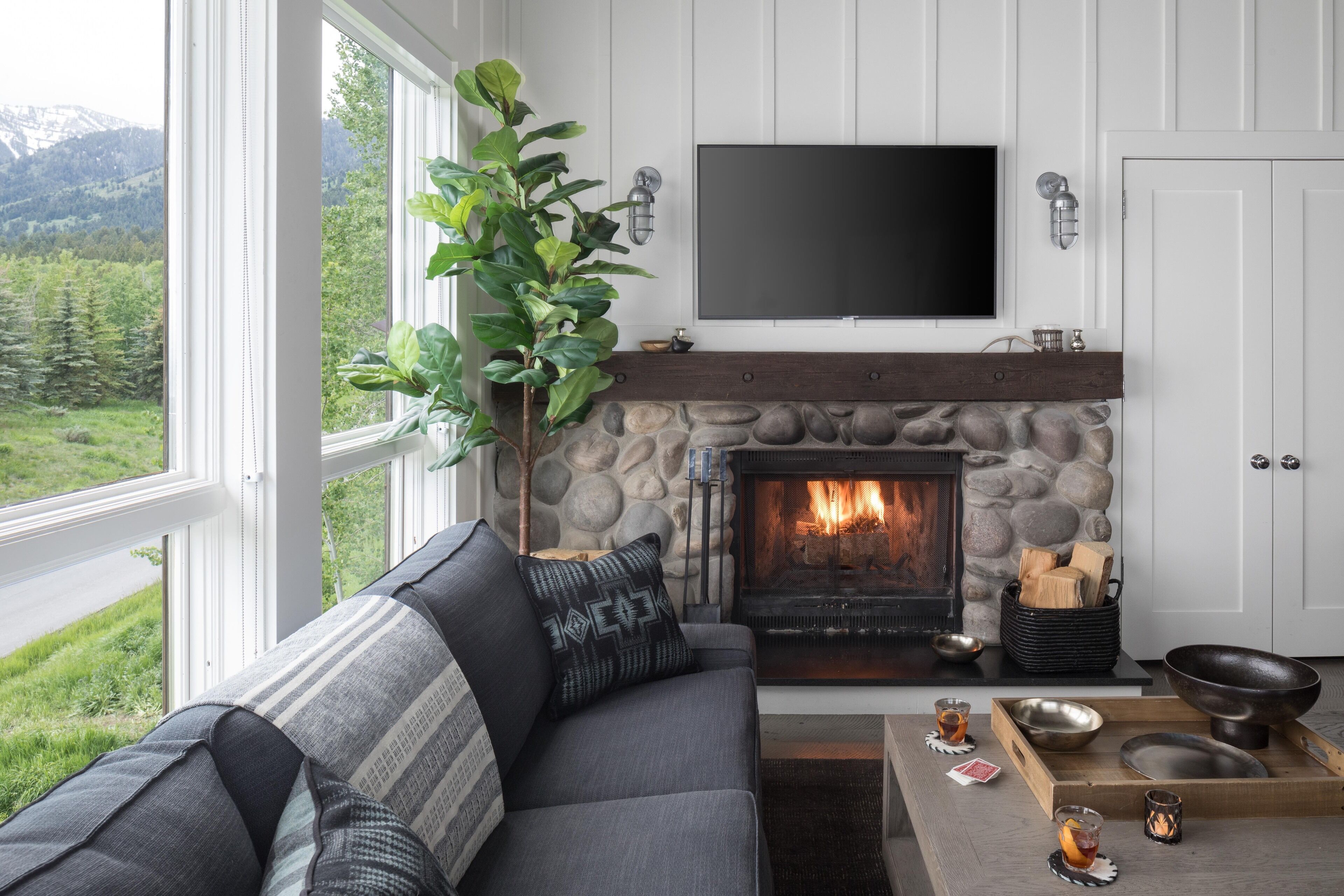 A living room with a stone fireplace, a gray sofa with patterned cushions, a flat-screen TV mounted above the fireplace, and a view of a green landscape through large windows.