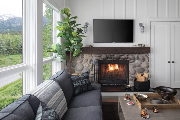 A living room with a stone fireplace, a gray sofa with patterned cushions, a flat-screen TV mounted above the fireplace, and a view of a green landscape through large windows.