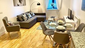 A modern living room with a grey sofa, a glass dining table, and a TV on a stand.