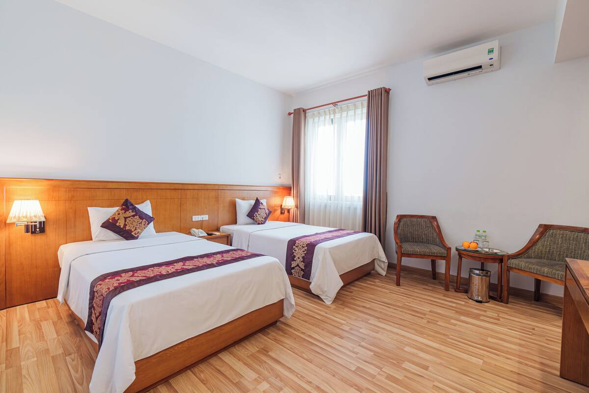 Une chambre d’hôtel avec deux lits, un bureau, une chaise, une poubelle et une fenêtre avec des rideaux.