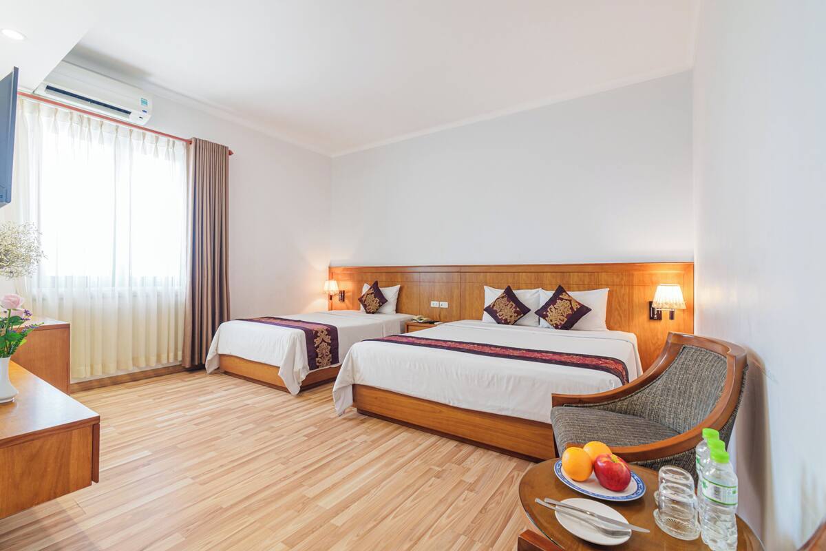 Une chambre d’hôtel avec deux lits, un plancher en bois, un bureau agrémenté d’un vase de fleurs et un plateau de petit-déjeuner avec des fruits et une bouteille d’eau.