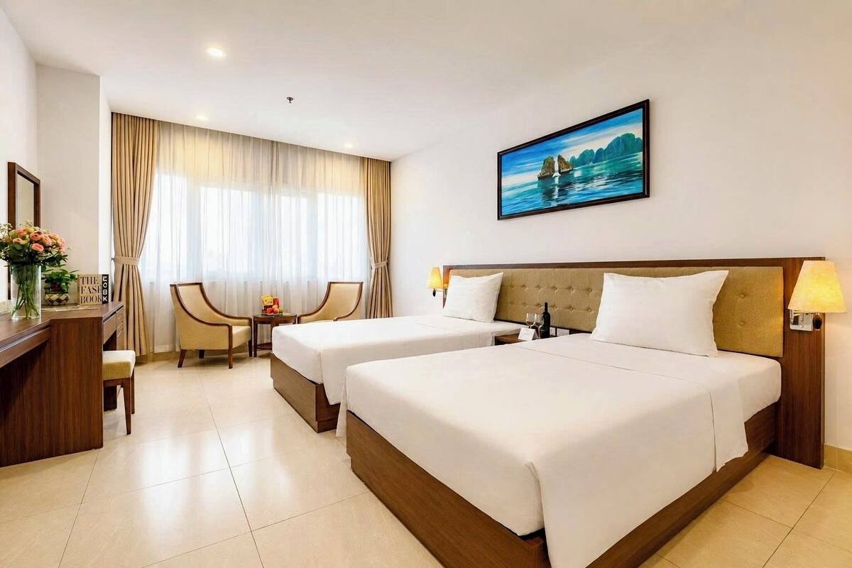 Une chambre d’hôtel avec deux lits, un bureau avec une chaise, une petite table avec des fruits, un vase avec des fleurs et un tableau au mur.