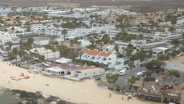 A coastal town with white buildings, a sandy beach, and a mountain in the background.