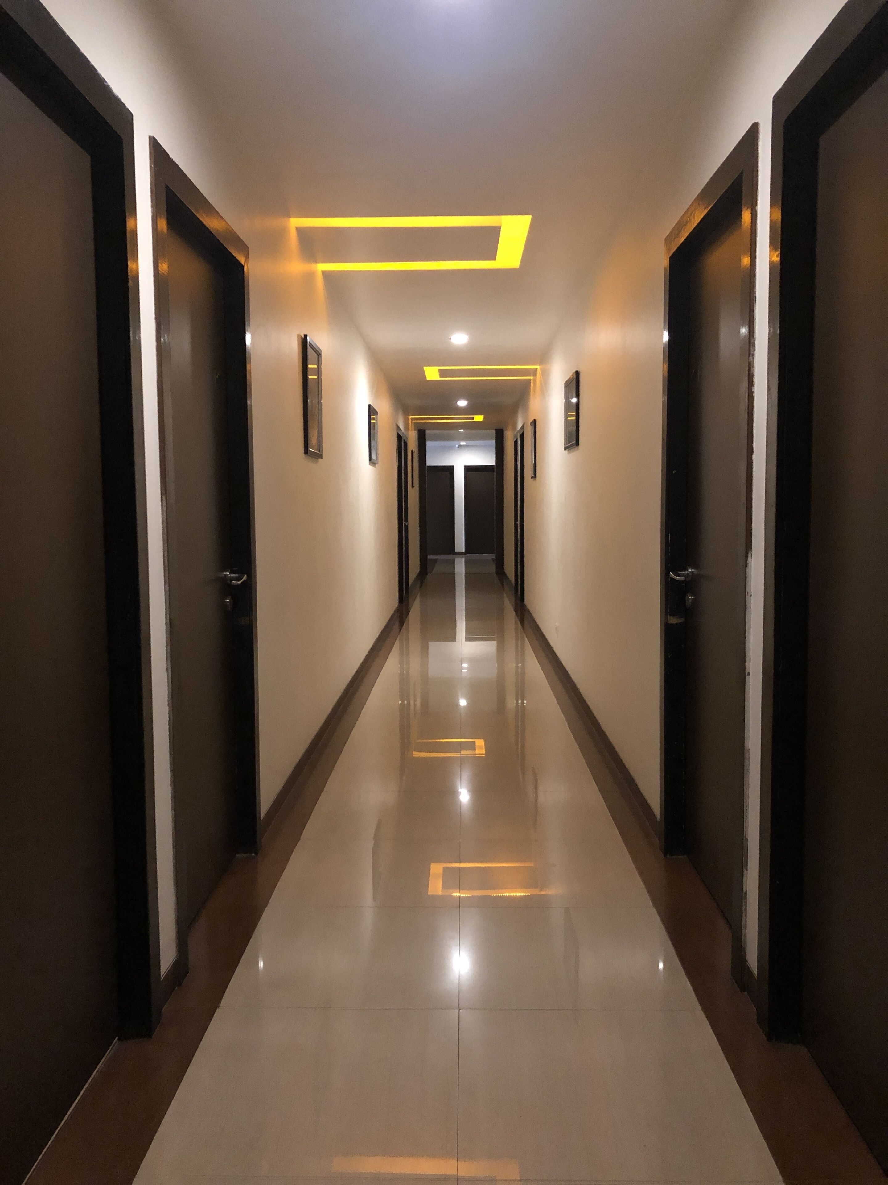 Un pasillo largo y estrecho con puertas a ambos lados, suelo de baldosas brillantes e iluminación empotrada en el techo.