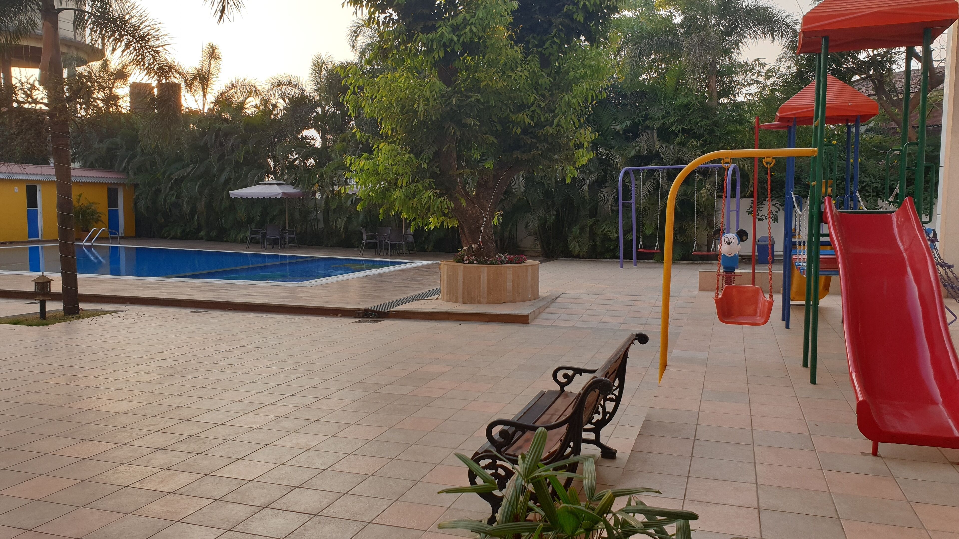 Zona de piscina con baldosas, una banca y un parque infantil con un tobogán rojo.