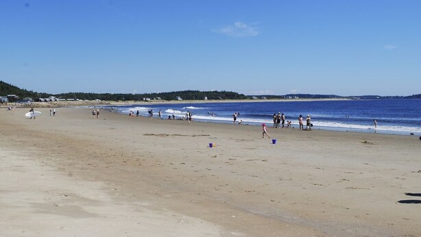 Una extensa playa de arena con gente caminando y jugando, cielo azul despejado y mar tranquilo.