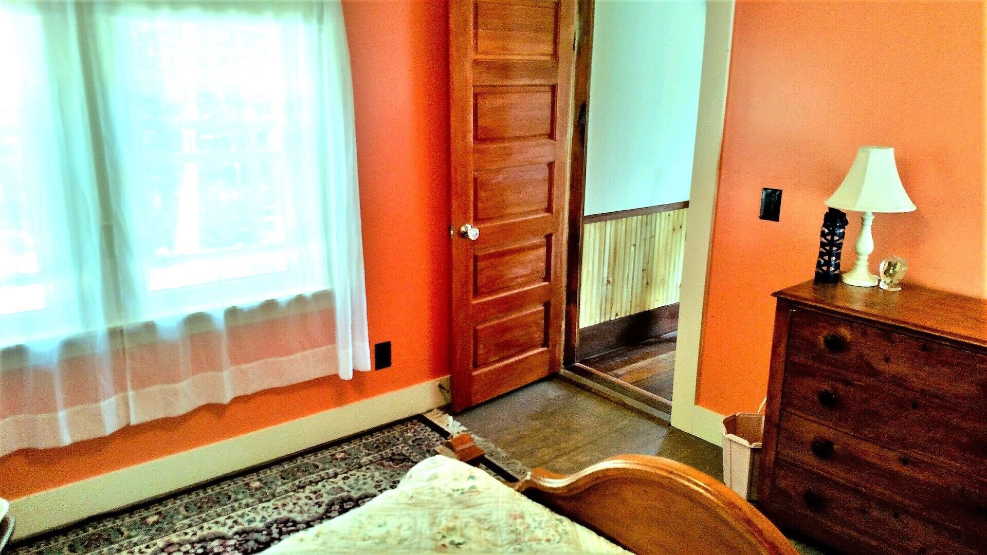 A room with a wooden door, a bed with a white bedspread, a dresser, and a lamp on the dresser.